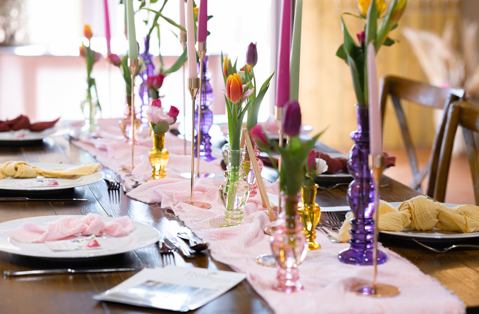 Ligne de vases en verre coloré et de chandeliers dorés sur un chemin rose pâle, tulipes et serviettes roses et ocre