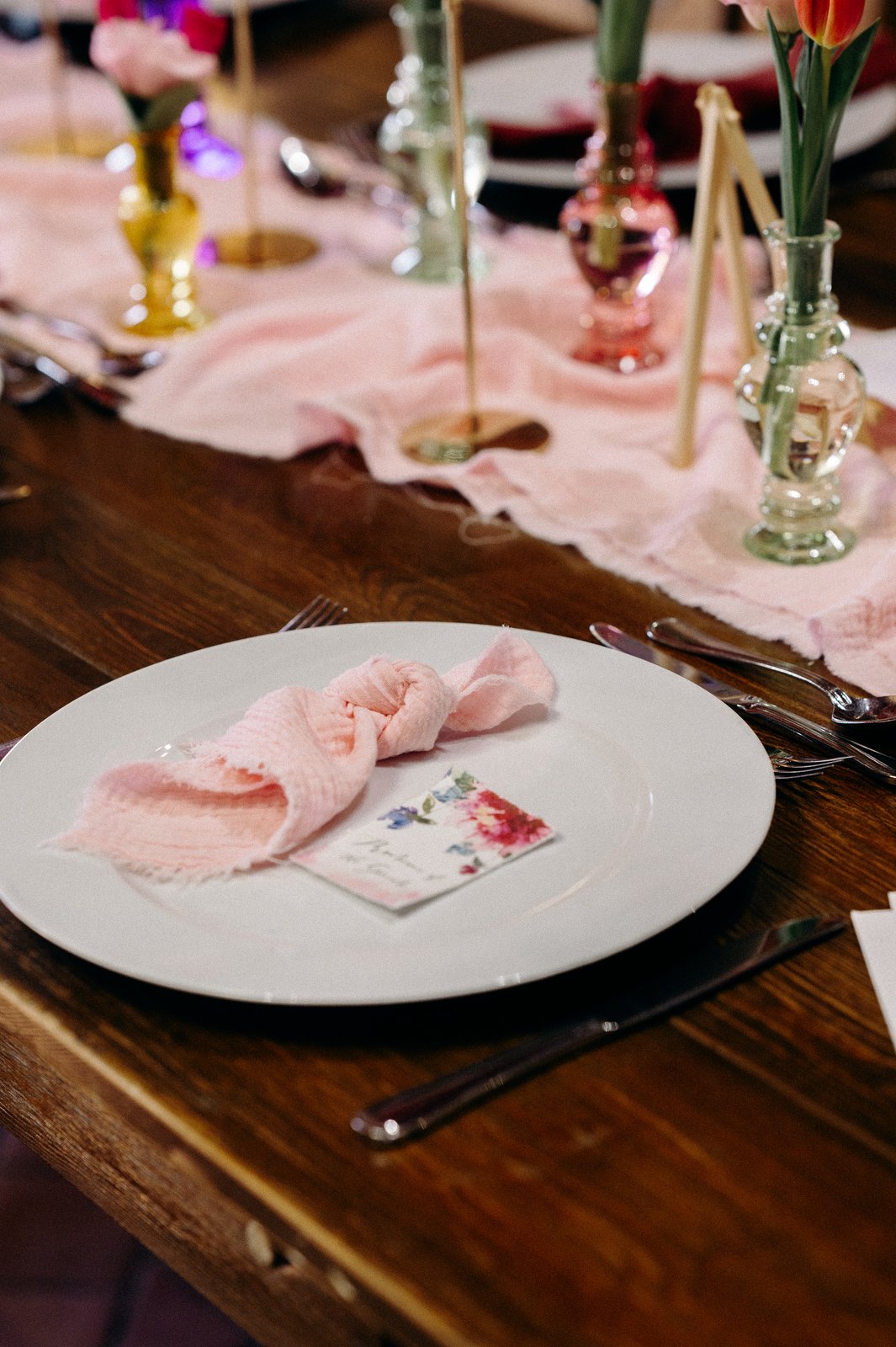 Assiette blanche, serviette rose froissée et marque-place fleuri sur une table en bois, vases en verre coloré derrière