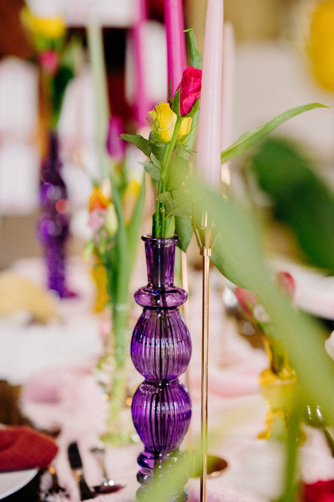Vase en verre violet strié garni de tulipes jaunes et rouges, bougies roses et vertes floues sur un chemin rose