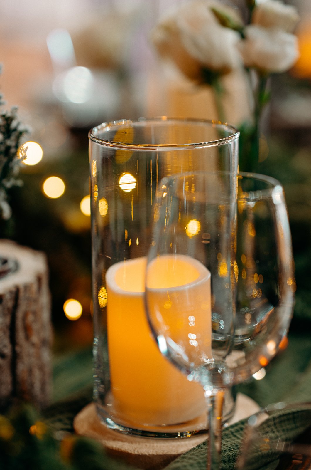 Bougie dans un cylindre en verre posé sur un rondin de bois, derrière un verre à pied, lumières floues en arrière-plan