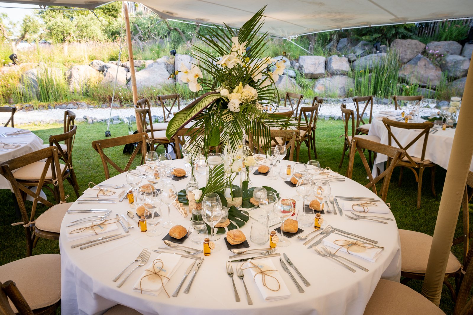 Table ronde nappée de blanc sous tente stretch, haut vase cylindre de palmes et d’orchidées, chaises bistrot à coussin