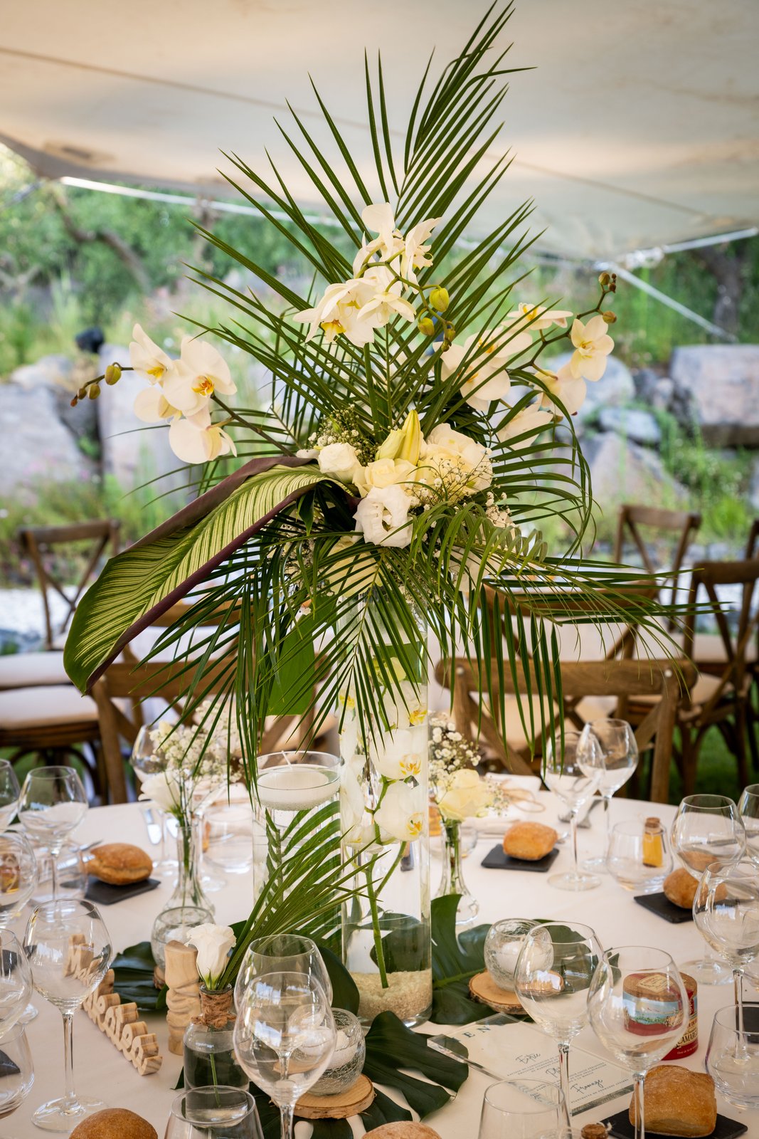 Centre de table tropical : palmes, orchidées et roses blanches dans un vase cylindre, feuilles de monstera sur la nappe