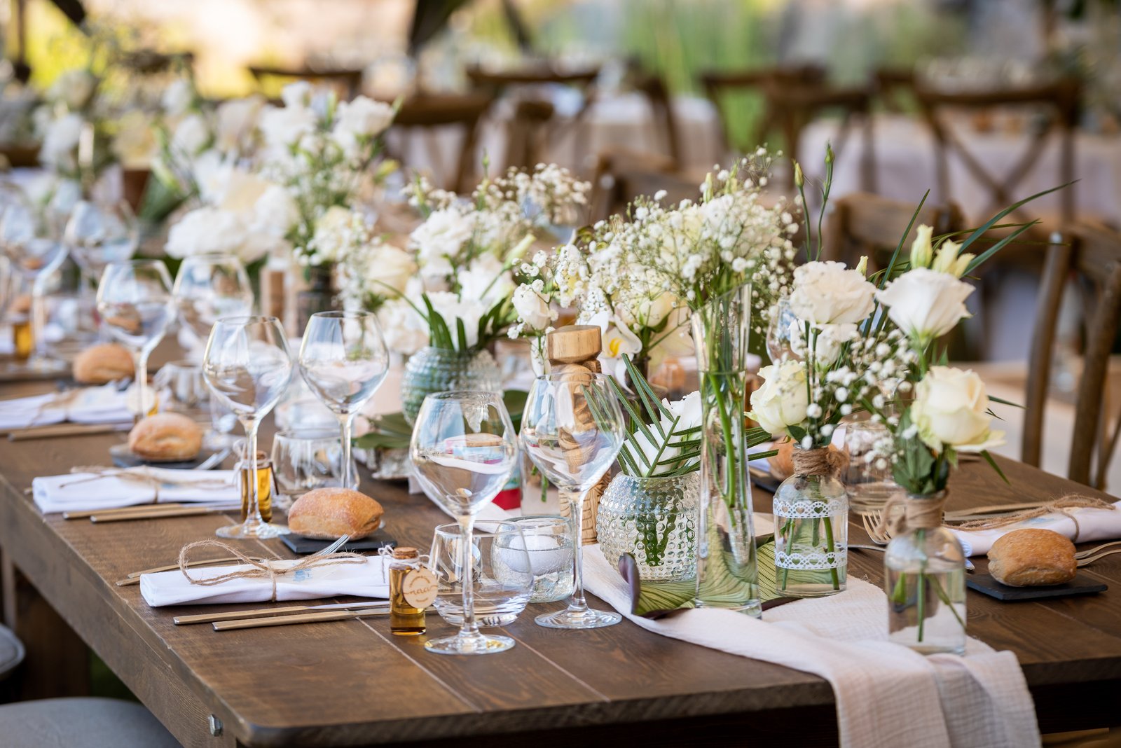 Table en bois dressée d’un chemin de table écru, soliflores de roses blanches et gypsophile, verres à pied et petits pains