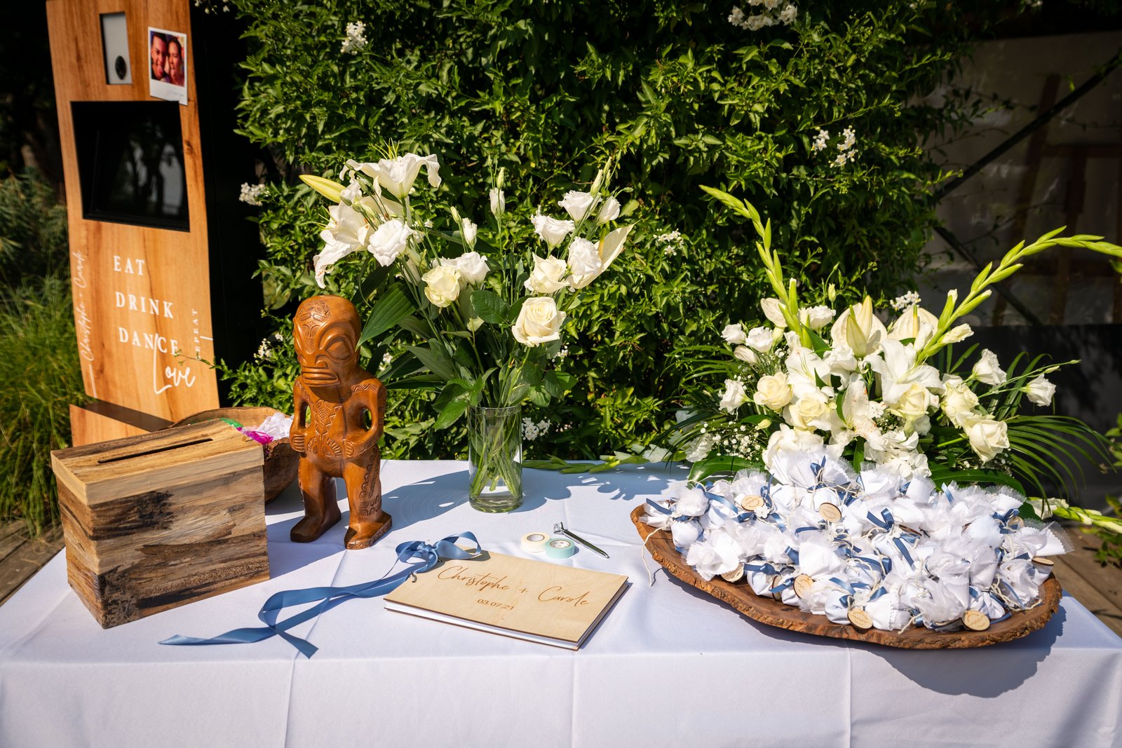 Table d’accueil nappée de blanc : borne photo en bois, urne en bois, livre d’or, bouquets blancs et dragées en tulle