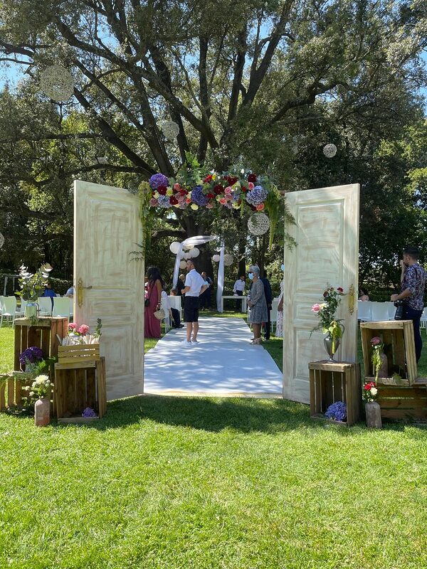 Portes de cérémonie blanches surmontées d’une guirlande de fleurs, caisses en bois fleuries et allée blanche dans un parc