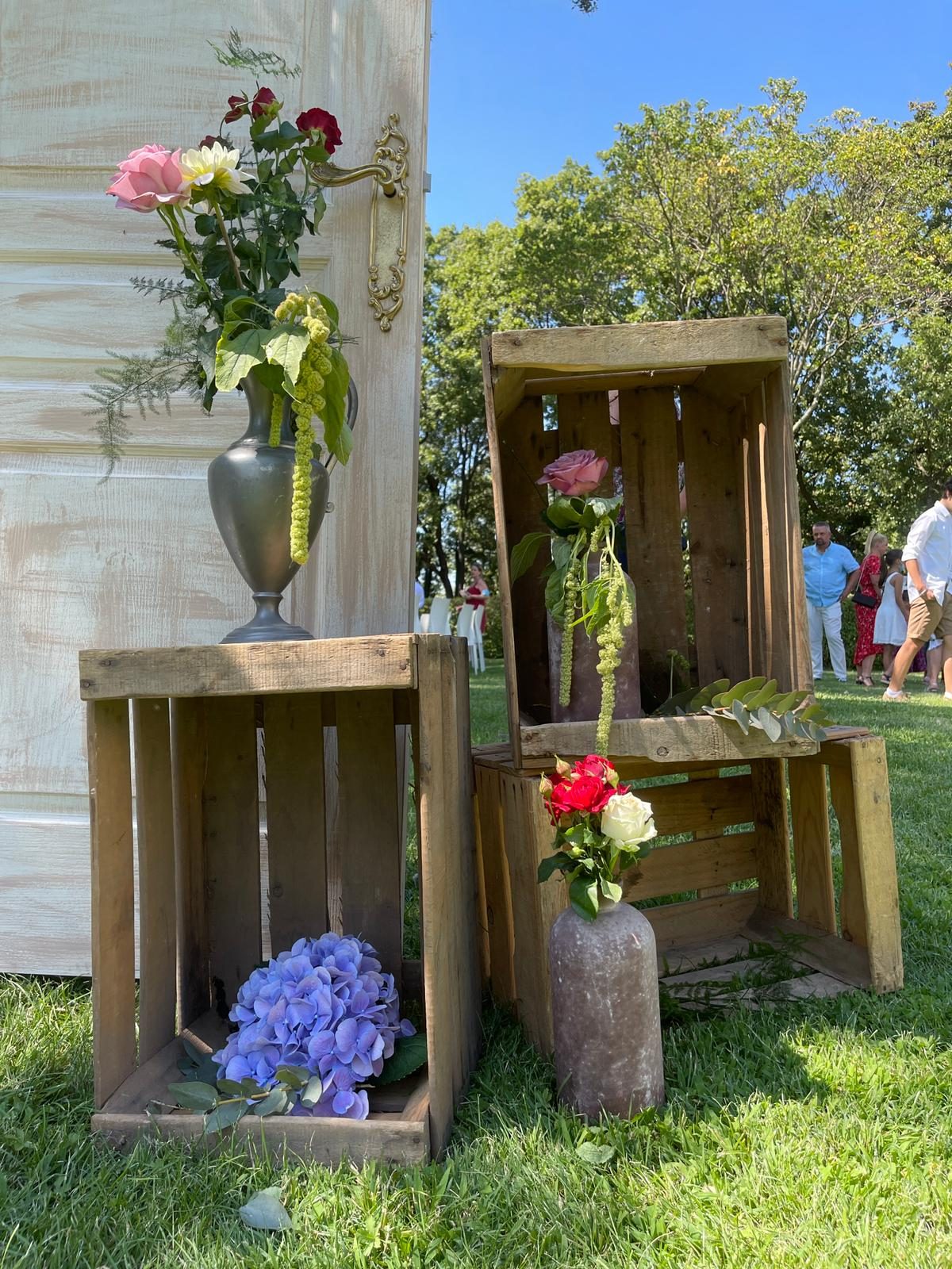 Caisses en bois empilées sur la pelouse avec hortensias bleus, roses et vase en étain devant une porte de cérémonie