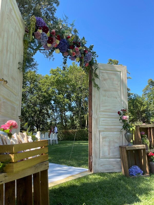 Porte de cérémonie ouverte sous une arche de fleurs colorées, caisses en bois fleuries sur la pelouse d’un domaine