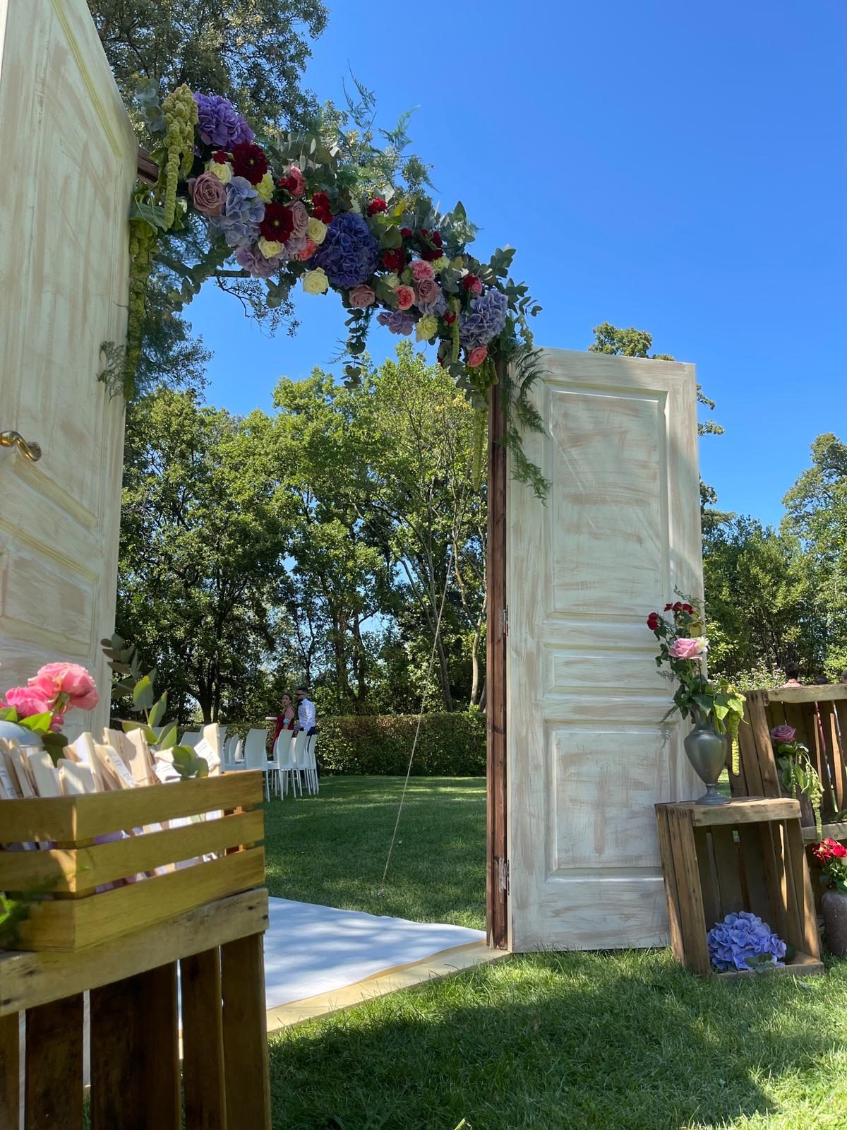Porte de cérémonie ouverte sous une arche de fleurs colorées, caisses en bois fleuries sur la pelouse d’un domaine