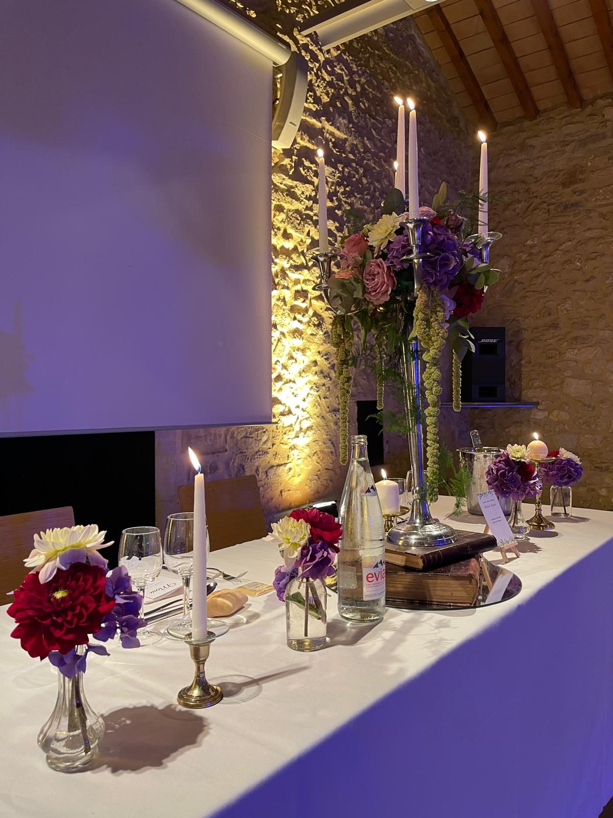Table d’honneur en nappe blanche, chandelier argenté fleuri posé sur des livres anciens, bougies et soliflores