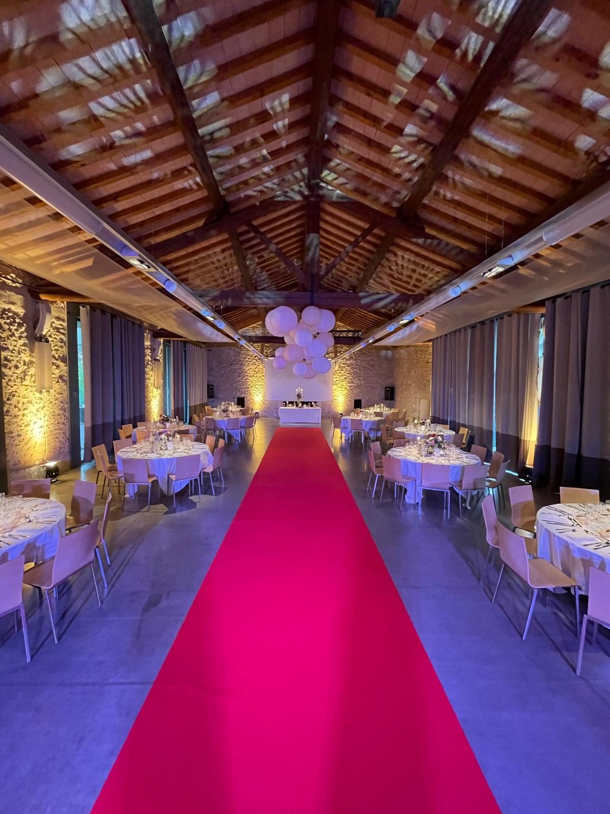 Salle de réception sous charpente en bois, tapis rouge central, tables rondes nappées et lanternes blanches suspendues