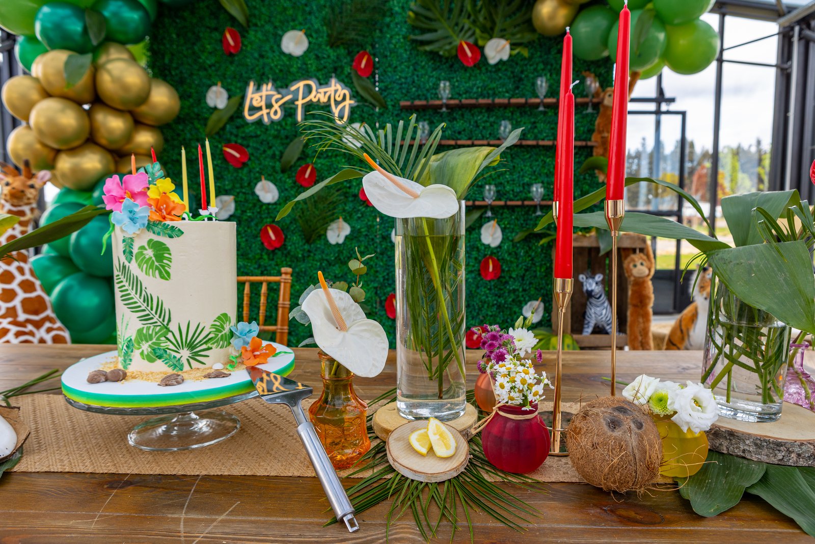 Gâteau aux feuilles tropicales, ananas, bougies rouges sur bougeoirs dorés et vases d’anthuriums devant un mur végétal