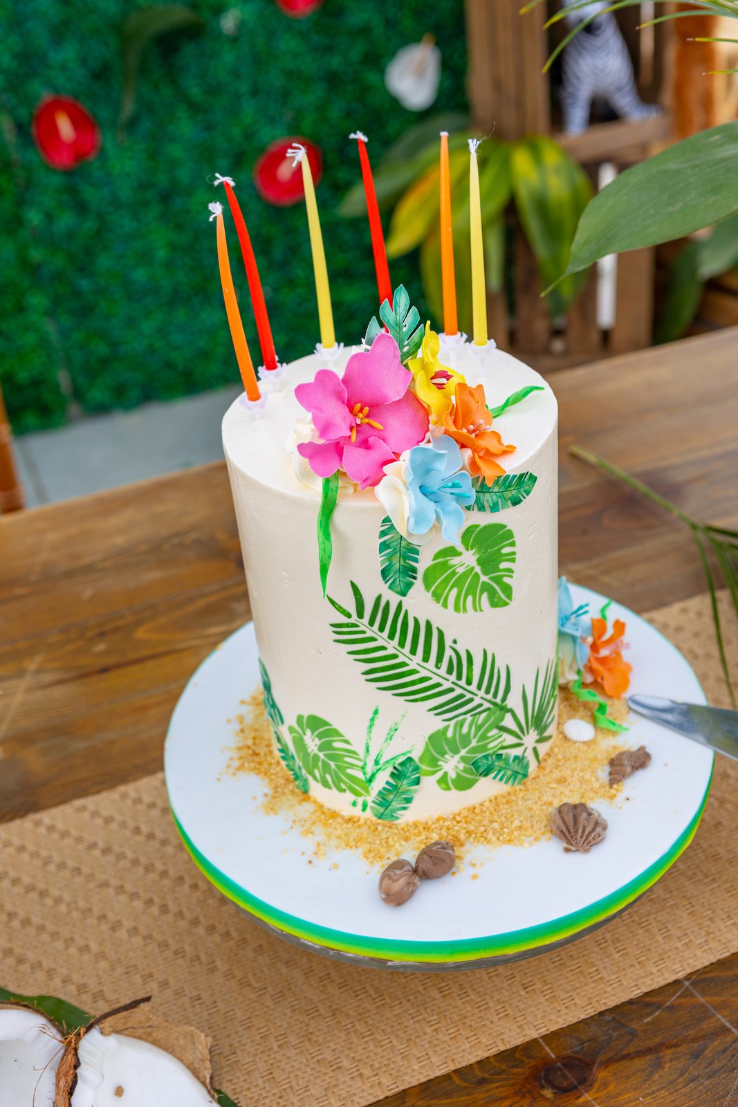 Gâteau blanc décoré de fleurs et feuilles tropicales, bougies colorées, posé sur un chemin de table tressé