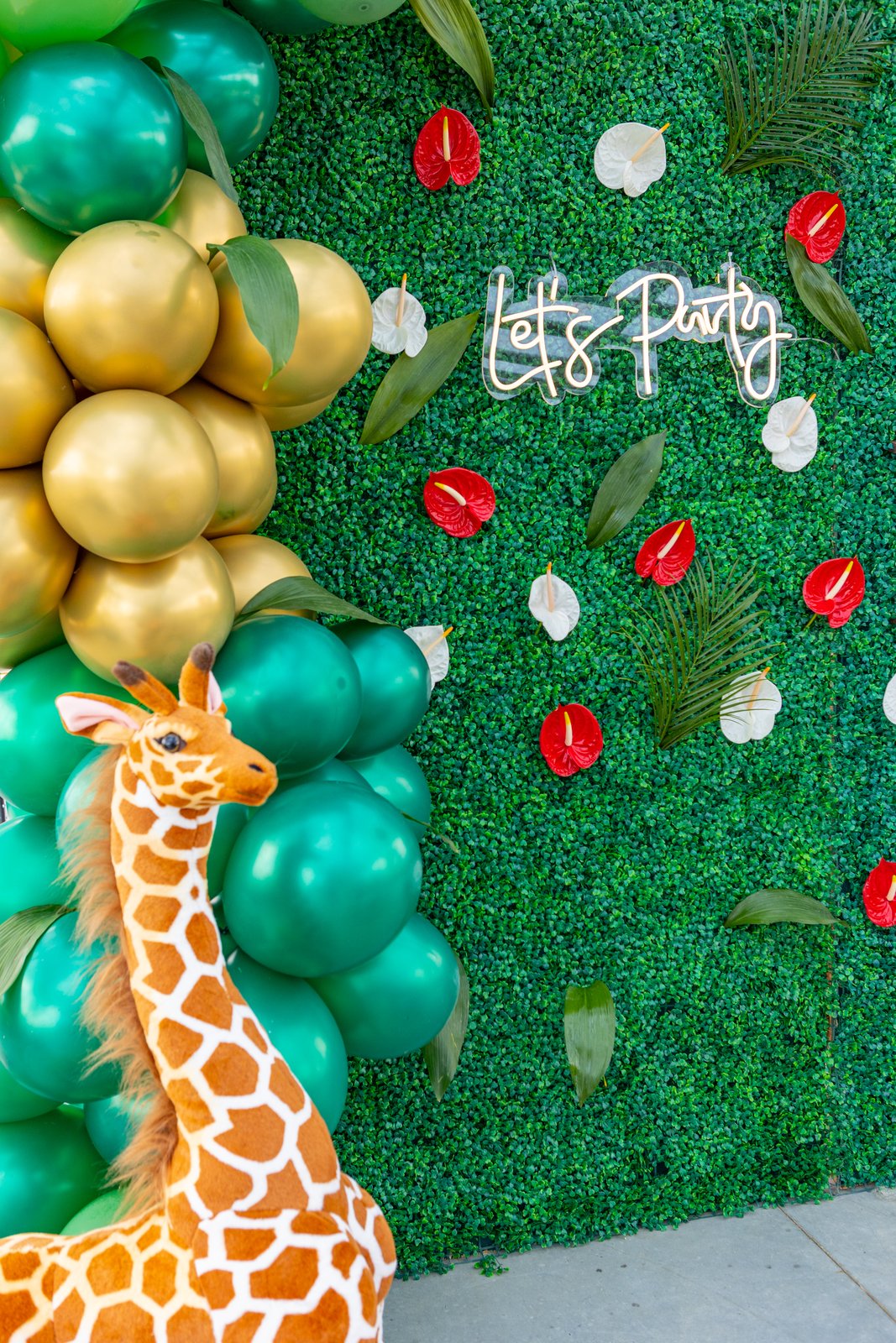 Néon « Let’s Party » sur un mur végétal à anthuriums, guirlande de ballons dorés et verts, girafe en peluche