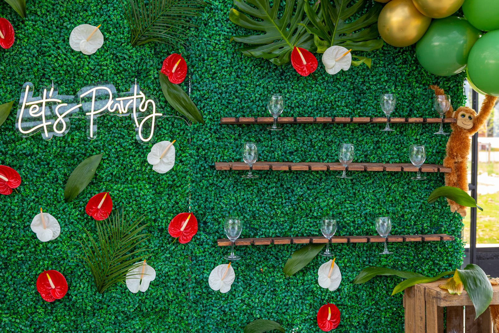 Étagères à flûtes sur un mur végétal semé d’anthuriums rouges et blancs, néon « Let’s Party » et ballons dorés