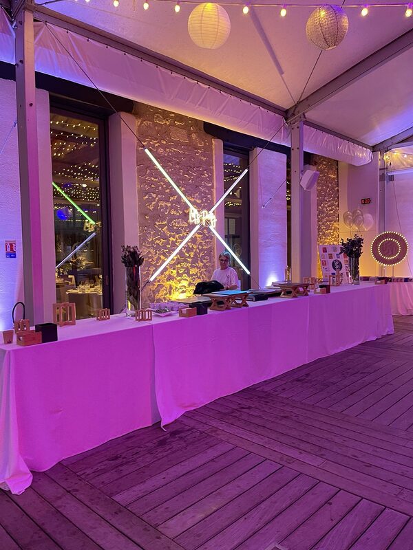 Long buffet nappé de blanc devant un mur de pierre éclairé, tubes lumineux croisés et néon « Let’s Party », guirlandes et lanternes au plafond