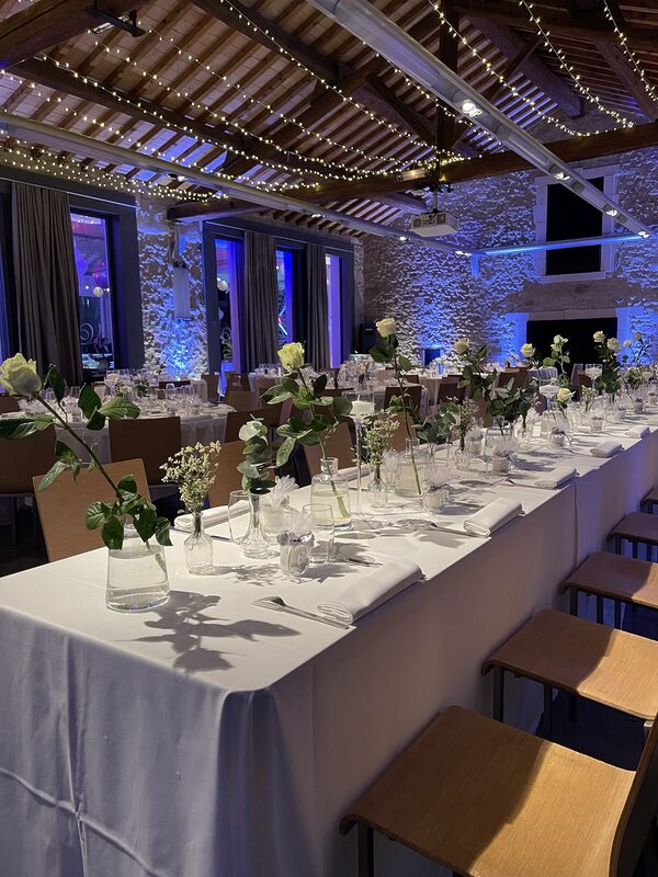 Longue table nappée de blanc dressée de soliflores, carafes et roses blanches, sous une charpente tendue de guirlandes lumineuses, au Domaine de Verchant