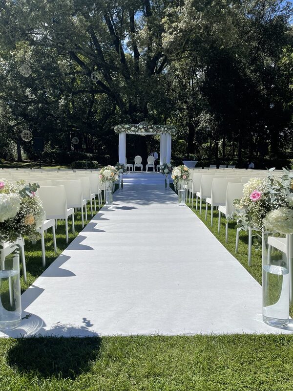 Allée de cérémonie au tapis blanc sous de grands arbres, chaises blanches, hauts vases cylindre fleuris et houppa drapée de voilage blanc couronnée de fleurs