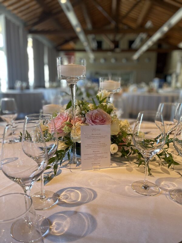 Centre de table de roses roses et blanches, bougies flottantes dans des vases sur pied et menu, sur une table ronde nappée de blanc