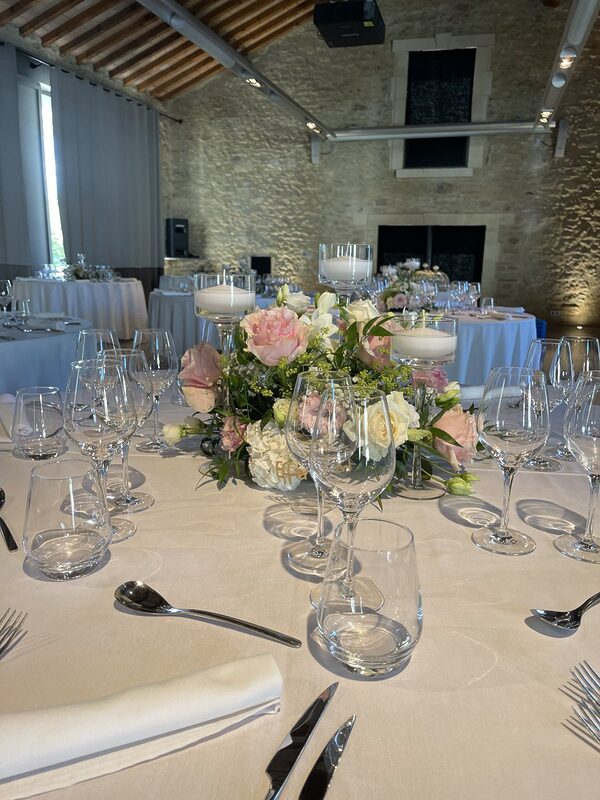 Tables rondes nappées de blanc dans une salle aux murs de pierre et charpente apparente, bouquets ronds et bougies flottantes sur pied