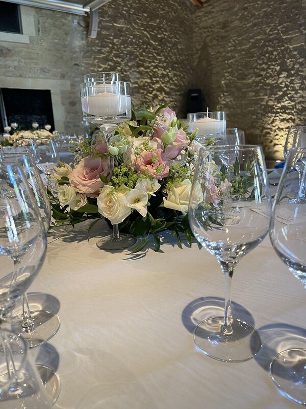 Bouquet bas de roses, lisianthus et alchémille entouré de bougies flottantes dans des vases sur pied, devant un mur de pierre éclairé