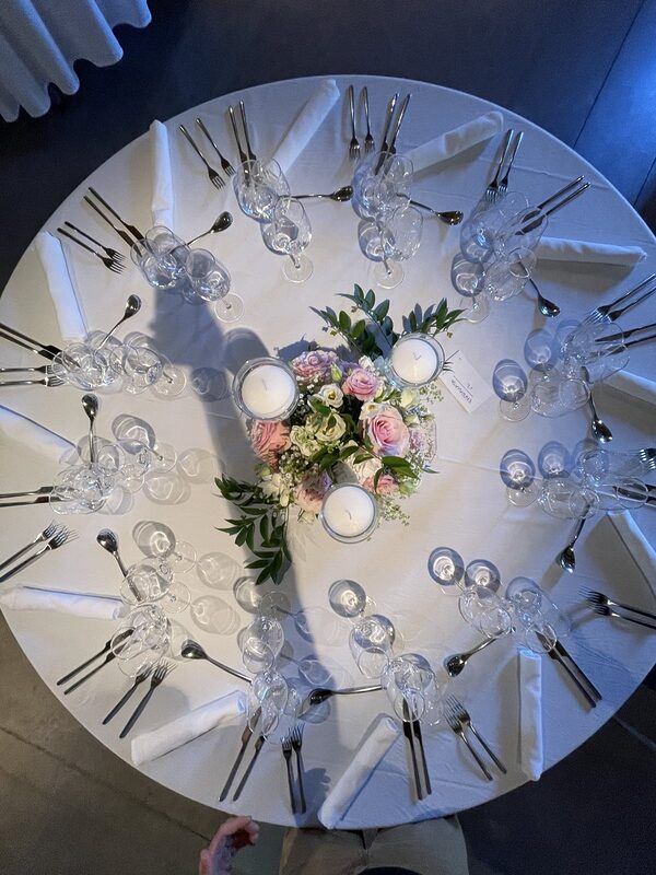 Table ronde vue du dessus : nappe blanche, couverts et verres disposés en cercle autour d’un bouquet et de trois bougies flottantes