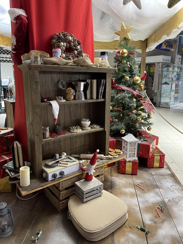Cuisine des lutins : buffet en bois garni de bocaux, moules et biscuits, petit lutin, sapin décoré et cadeaux rouges sur un plancher saupoudré de neige