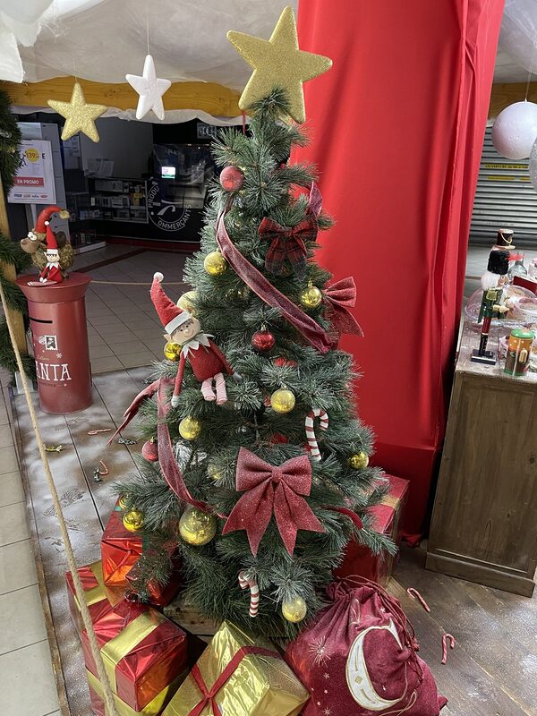 Sapin décoré de boules or et rouges, de nœuds pailletés et d’un lutin, boîte aux lettres rouge et casse-noisette à côté, sous un ciel de voilage