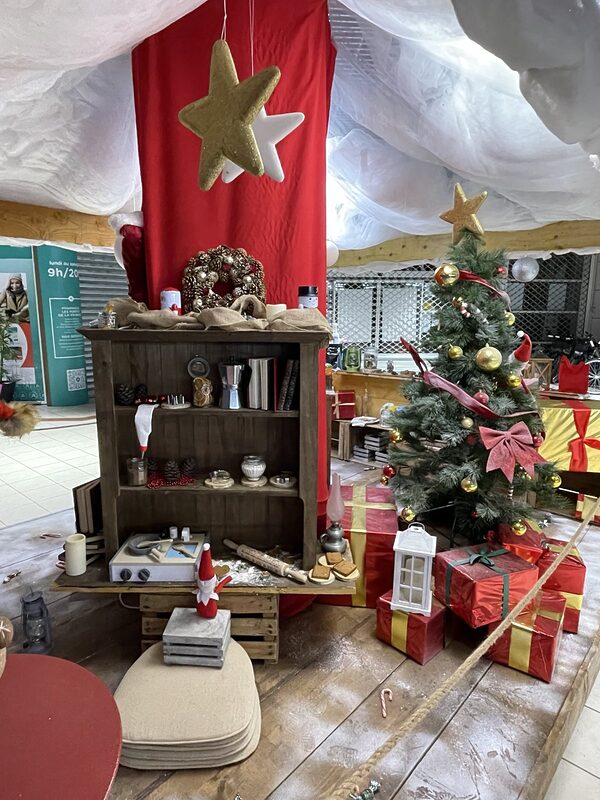 Vue d’ensemble de la cuisine des lutins sous un voilage blanc et des étoiles suspendues, pilier habillé de rouge, sapin et cadeaux