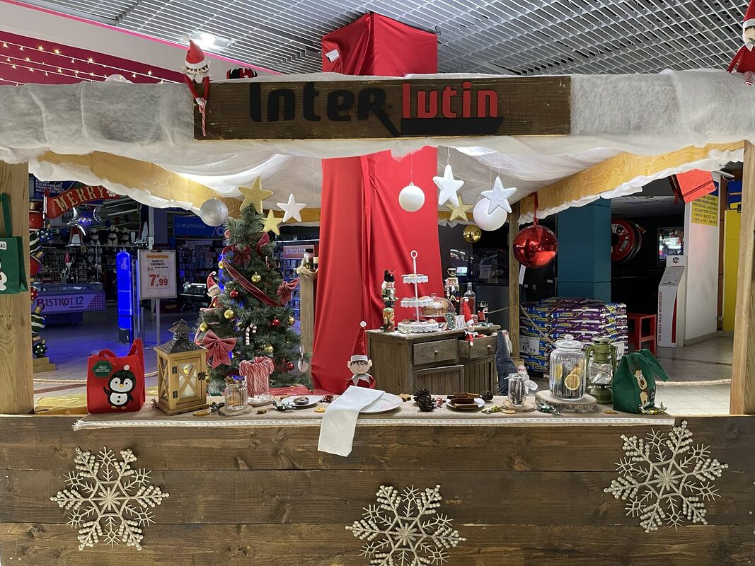 Chalet en bois « Inter lutin » au toit enneigé, comptoir garni de bocaux, lanterne, sapin décoré et lutins assis sur le toit