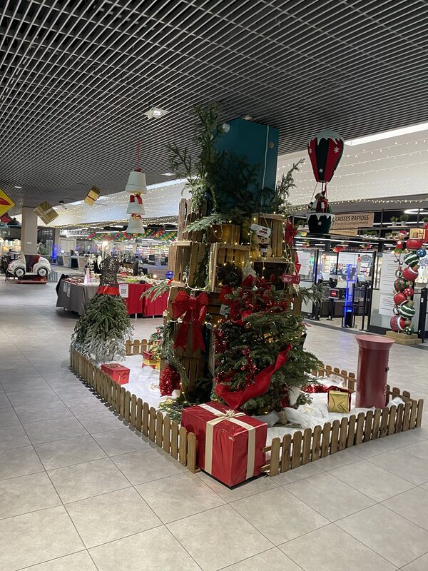 Îlot de Noël autour d’un pilier : cagettes en bois et sapins naturels, gros nœuds rouges, cadeaux et boîte aux lettres, barrière en bois