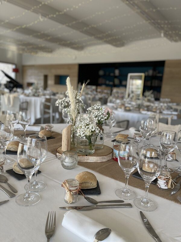 Centre de table champêtre sur rondin de bouleau : gypsophile, pampas et roses poudrées en bocaux, photophore craquelé, chemin de jute sur nappe blanche
