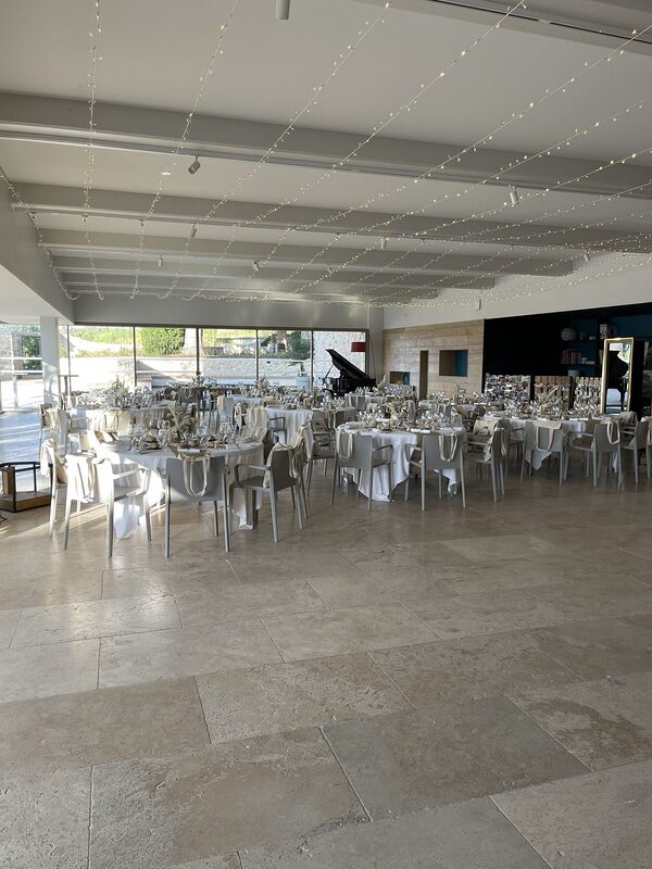 Salle de réception lumineuse sous un plafond de guirlandes LED tendues, tables rondes nappées de blanc aux chemins de jute