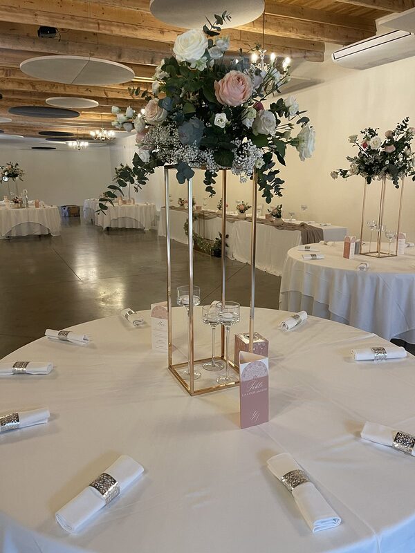Salle de réception sous charpente en bois : haute structure dorée couronnée de roses blanches et poudrées, bougies flottantes sur pied, tables rondes nappées de blanc