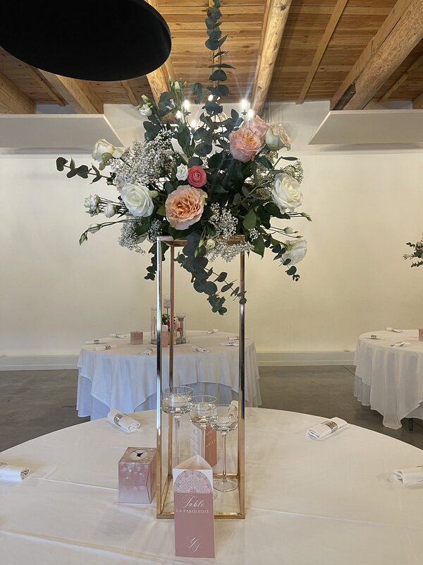 Centre de table haut : structure dorée portant un bouquet de roses pêche et blanches, gypsophile et eucalyptus, bougies flottantes sur pied