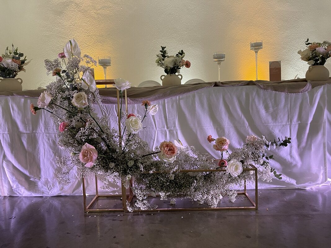 Composition basse de gypsophile, roses et anthuriums blancs sur structures dorées au pied de la table d’honneur, chemin en gaze taupe et vases en terre