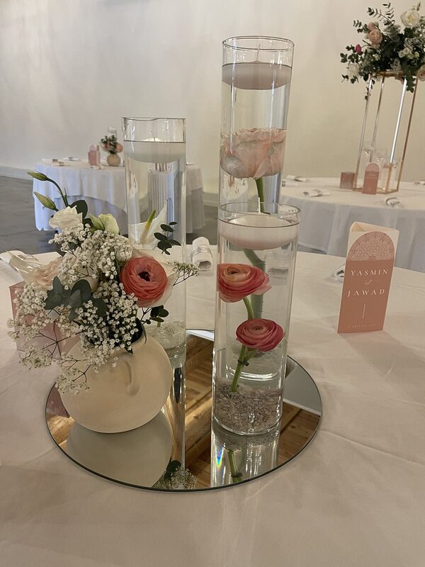 Vases cylindres aux renoncules immergées et bougies flottantes sur miroir rond, petit vase en terre à anses garni de roses et gypsophile